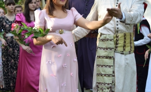 Zdjęcie przedstawia uczestników wydarzenia. Na pierwszym planie para – kobieta w różowej sukni, trzymająca w ręce kwiaty oraz mężczyzna w białym stroju, przepasany ozdobnym, żółtym pasem.
