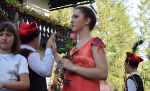 Zdjęcie przedstawia uczestników wydarzenia. Na pierwszym planie stoi młoda kobieta ubrana w czerwoną suknię. W dłoni trzyma różę.