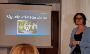 Zdjęcie przedstawia ekran sufitowy, na którym widoczny jest slajd prezentacji o tytule Ogrody w świecie islamu. Po prawej stronie znajduje się kobieta.