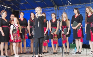 Zdjęcie przedstawia uczestników wydarzenia, którzy znajdują się na scenie. Kobiety są ubrane w czarne sukienki, a w pasie mają czerwoną w stylu ludowym szarfę. Tworzą półkole, w którego centrum śpiewa jedna z nich.