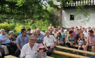 Zdjęcie przedstawia uczestników wystawy, którzy siedzą na ławkach w plenerze. Otacza ich bujna roślinność z lewej strony. Jest słoneczny dzień.