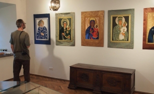Zdjęcie przedstawia jednego z uczestników wystawy, który przygląda się obrazom wiszącym na ścianie. Znajduje się po lewej stronie zdjęcia, a jego cień pada na fragment obrazu.