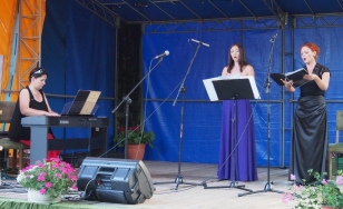 Zdjęcie przedstawia kobiety występujące na scenie. Śpiewają one do mikrofonów. Po lewej stronie na pianinie elektrycznym gra kobieta.