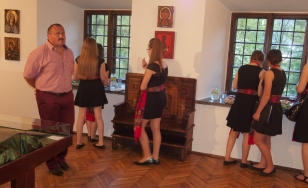 Zdjęcie przedstawia uczestników spotkania, którzy korzystają z poczęstunku w jednym z pomieszczeń dworu. Kobiety ubrane są w czarne sukienki. W pasie posiadają folklorystyczne szarfy.