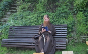 Zdjęcie wykonane w plenerze przedstawia zakonnika czytającego książkę. Siedzi on na drewnianej ławce w kolorze szarym. Znajduje się w otoczeniu bujnej roślinności.