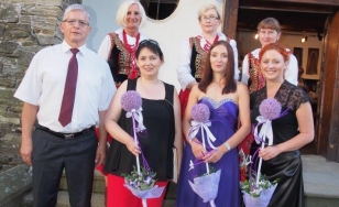 Zdjęcie grupowe uczestników wystawy na tle dworu. Na pierwszym planie mężczyzna i trzy kobiety z fioletowymi kwiatami. W drugim rzędzie kobiety w tradycyjnych strojach ludowych.