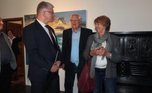 Zdjęcie przedstawia uczestników wystawy. Na tle wielkoformatowego obrazu olejnego stoją dwaj mężczyźni i jedna kobieta. Wszyscy są radośni.