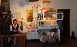 Zdjęcie przedstawia uczestniczkę wystawy na tle ekspozycji. Na białej komodzie ustawione są kwiatowe aranżacje oraz figurki w stylu rustykalnym.