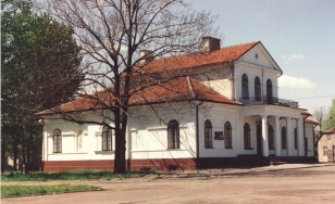 Zdjęcie przedstawia dworek w Wadowicach widziany z boku. Budynek ma białą fasadę i czerwony, spadzisty dach. Nad wejściem do dworu znajduje się taras podtrzymywany kolumnami.