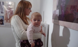 Zdjęcie przedstawia uczestników wystawy stojących na tle ekspozycji – matka trzymająca dziecko na rękach patrzy na obraz.