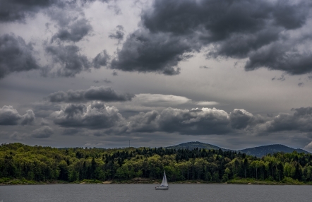 krajobraz z widokiem na jezioro i góry; pośrodku żaglówka, w tle porośnięte lasem pagórki; na niebie chmury
