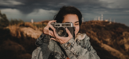 dziewczyna trzymająca przy oku aparat fotograficzny, w tle zalesione pagórki i zachmurzone niebo