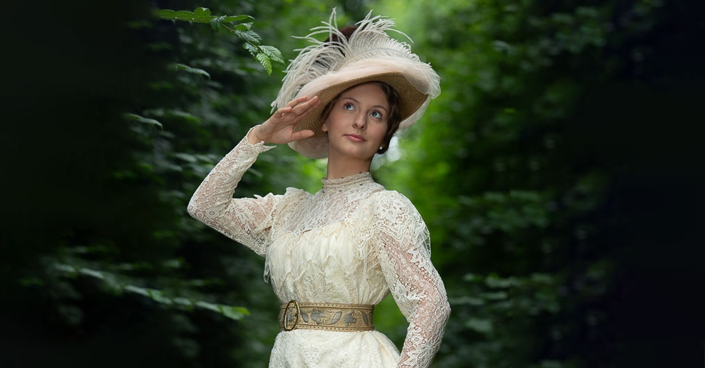 zdjęcie pięknej młodej kobiety,  w białej koronkowej sukni, na głowie ma biały kapelusz z piórami, w  tle zieleń lasu
