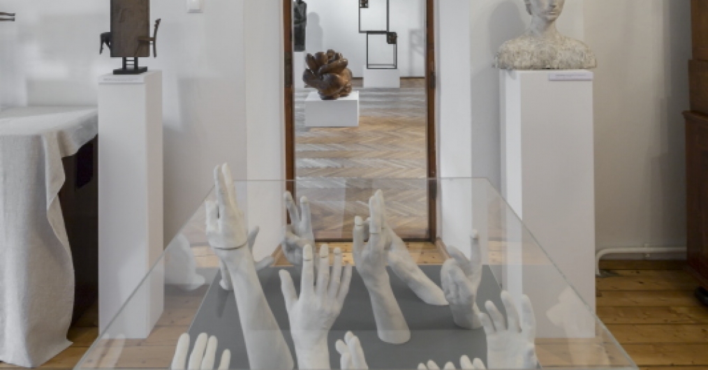 Zdjęcie przedstawia część wystawy. Ujęcie zogniskowane na gablocie z rzeźbami dłoni. W głębi znajduje się przejście do kolejnego pomieszczenia. Po jego prawej stronie znajduje się popiersie kobiece.