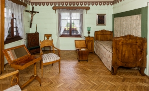 Zdjęcie przedstawia sypialnię. Ściany są w kolorze zielonym. Po prawej stronie stoi zabytkowe drewniane łóżko, nad którym wisi obraz z Matką Boską. Po lewej stronie w kącie wisi krucyfiks. W środkowej części jest okno.