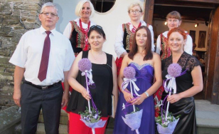 Zdjęcie grupowe uczestników wystawy na tle dworu. Na pierwszym planie mężczyzna i trzy kobiety z fioletowymi kwiatami. W drugim rzędzie kobiety w tradycyjnych strojach ludowych.