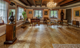 Zdjęcie przedstawia salon. Po lewej stronie wyeksponowane jest zabytkowe naczynie. W głębi otwarty fortepian. Po prawej stronie w kącie zegar stojący. Na ścianach portrety i obrazy. Znajdują się dwa krzesła przy stoliku.