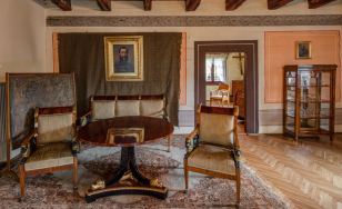 Zdjęcie przedstawia salon. Na pierwszym planie znajduje się stół, przy nim zabytkowe krzesła i sofa. Meble stoją na dywanie. Na ścianie wisi obraz na tle dywanu. Po prawej stronie są otwarte drzwi do kolejnego pomieszczenia.