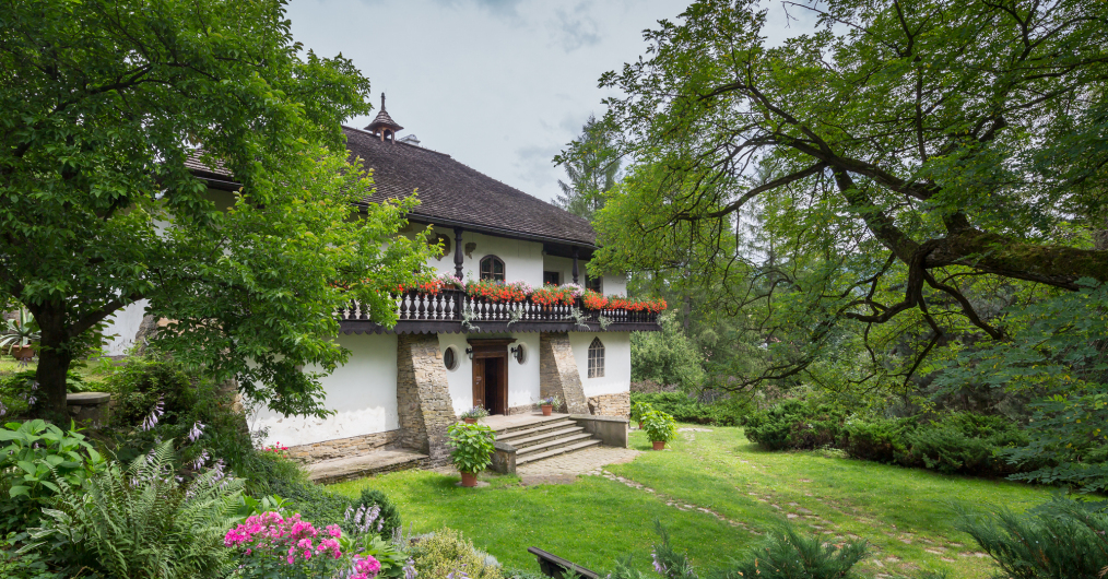 Widok wejścia do zabytkowego dworu, otoczonego zielonym ogrodem z rabatą różowych kwiatów.